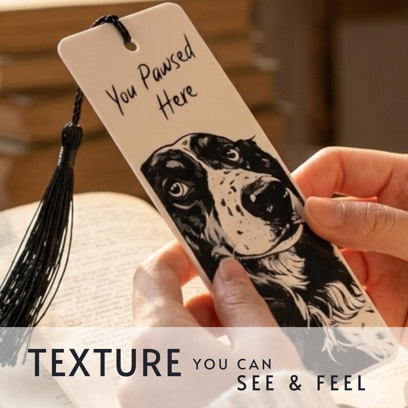 May include: A rectangular off-white bookmark featuring a black and white illustration of a dog's face and the text "You Pawsed Here" printed in black. The bookmark includes a hole at the top with an attached black decorative tassel.
