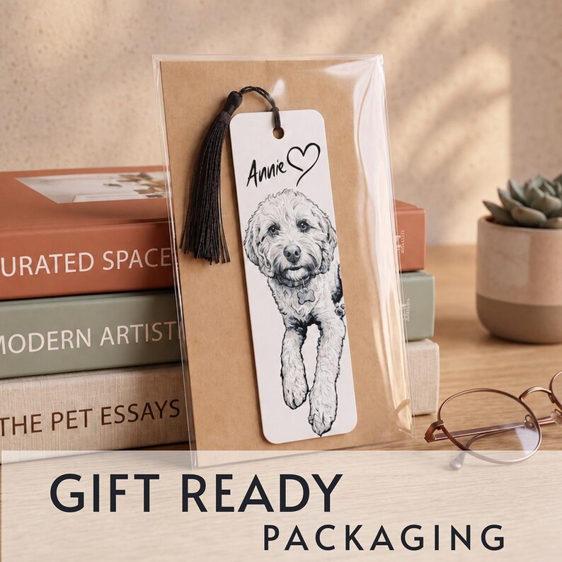 May include: A white rectangular bookmark featuring a black and white sketch of a fluffy dog, the name Annie, and a heart symbol. The bookmark includes a black tassel at the top and is enclosed in clear plastic packaging against a brown card backing.