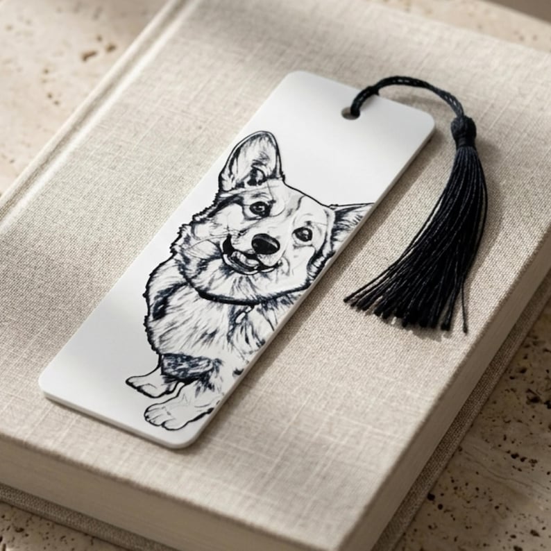 May include: A rectangular white bookmark featuring a detailed black and white line drawing of a Corgi dog. The bookmark has a small hole at the top with a black thread tassel attached, creating a functional accessory for marking pages in a book.