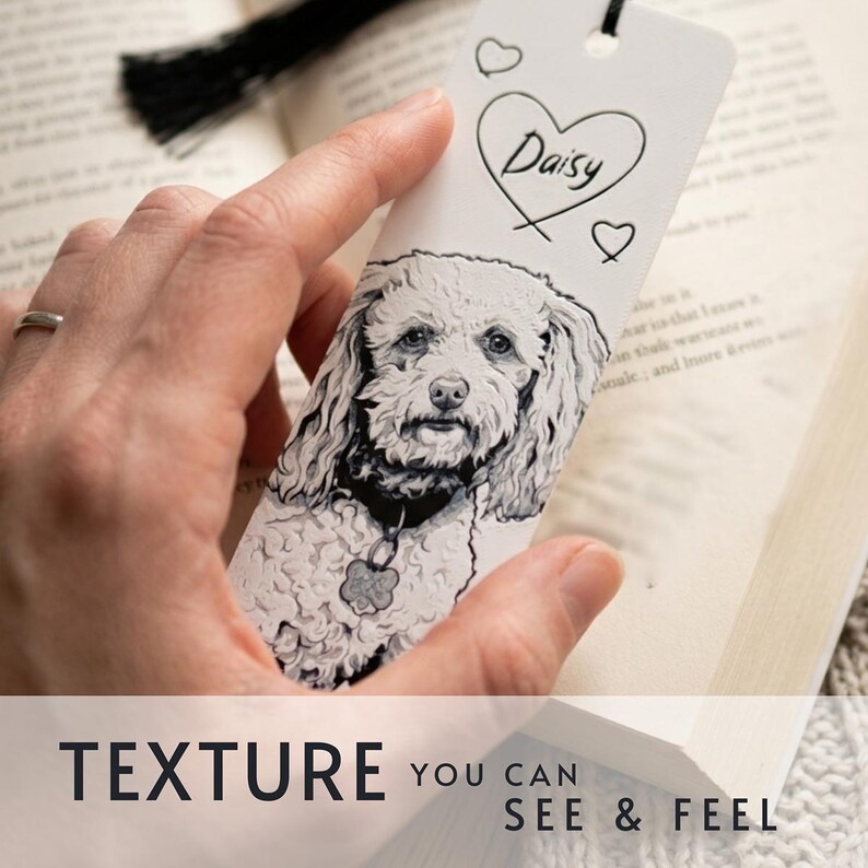 May include: A white rectangular bookmark featuring a detailed black and white illustration of a curly-haired dog wearing a collar with a tag. The top of the bookmark displays the name Daisy written inside a heart outline with two smaller hearts.