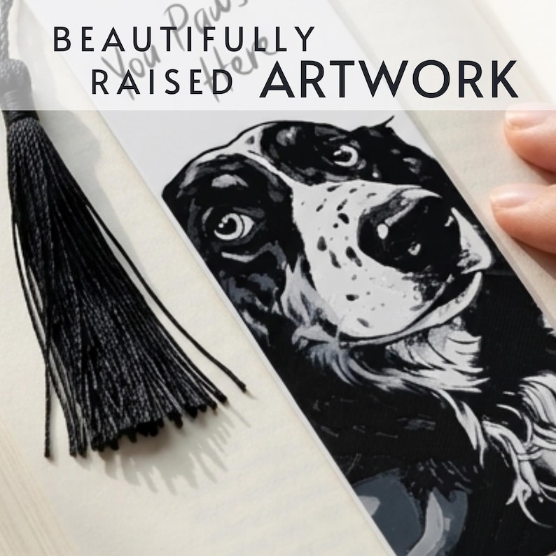May include: A white rectangular bookmark featuring a black and white illustration of a dog's face with a black tassel attached to the top. The bookmark displays the text BEAUTIFULLY RAISED ARTWORK at the top and You Paws-itively better below.