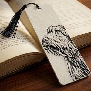 May include: A rectangular off-white bookmark featuring a detailed, embossed black illustration of a shaggy-haired dog's head in profile. The bookmark includes a hole at the top with a thin black tassel attached, resting against an open book.