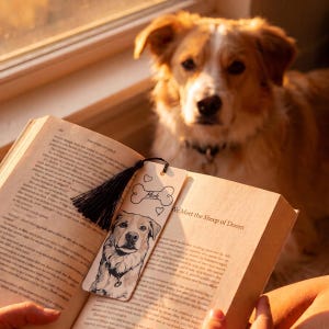 May include: A rectangular wooden bookmark featuring a black ink illustration of a dog, a bone-shaped graphic with the name "Merle" written inside, and small heart outlines. A black tassel is attached to the top of the bookmark.
