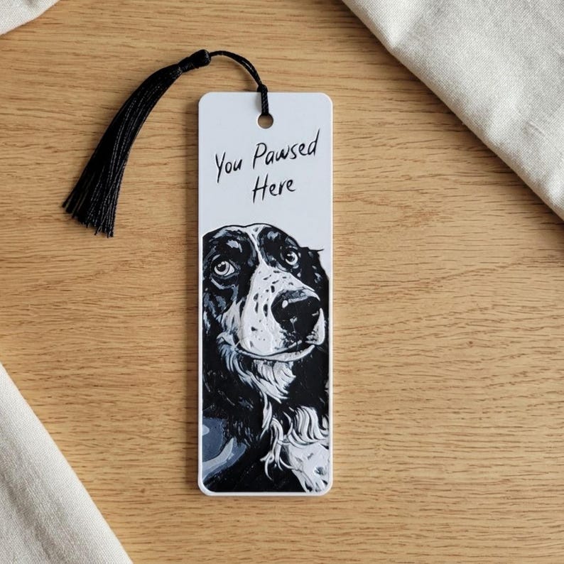 May include: A rectangular white bookmark featuring a black and white illustration of a dog's face. The text "You Pawsed Here" is printed in black ink above the dog. A black silk tassel is attached to the top through a circular hole.