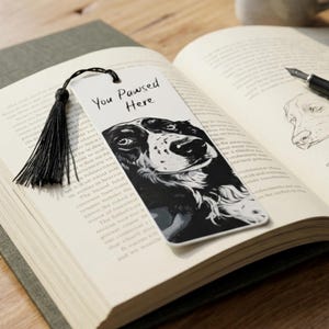 May include: A rectangular white bookmark featuring a black and white illustration of a dog's face and the text You Pawsed Here in black script. A black thread tassel is attached to the top of the bookmark through a small circular hole.