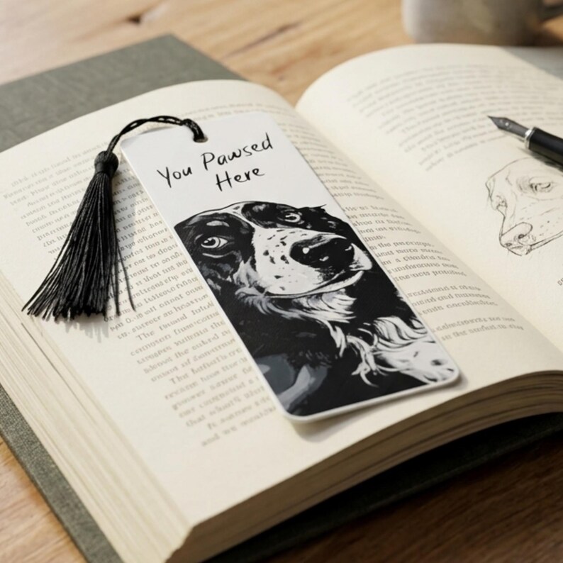 May include: A rectangular white bookmark featuring a black and white illustration of a dog's face and the text You Pawsed Here in black script. A black thread tassel is attached to the top of the bookmark through a small circular hole.