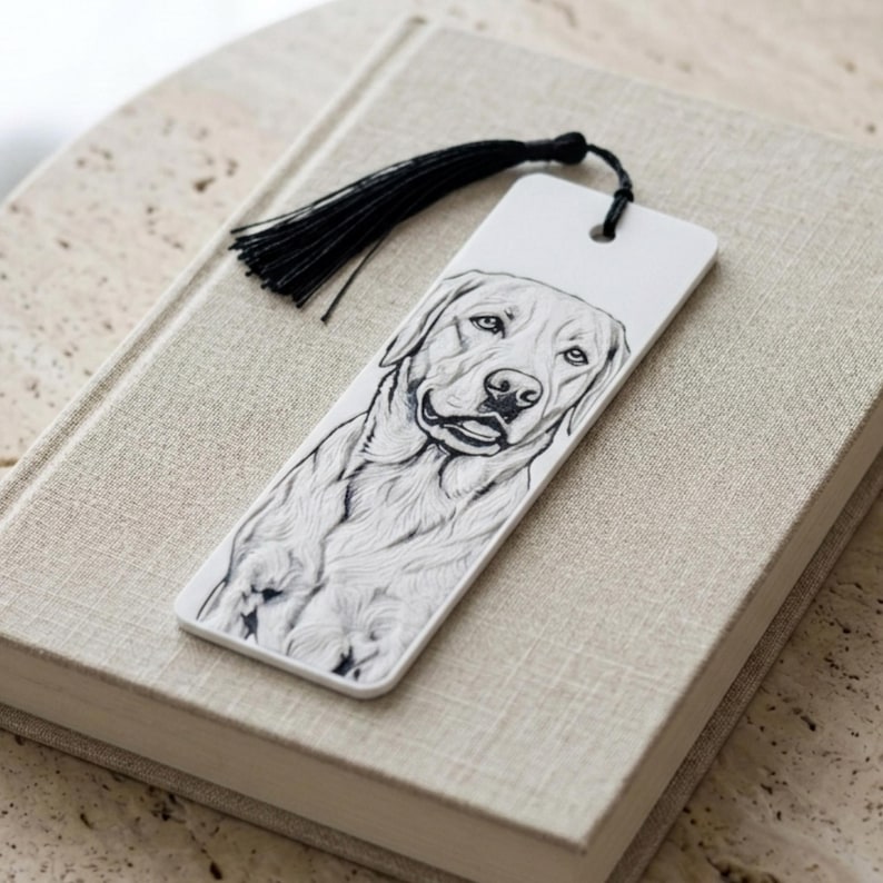 May include: A rectangular white bookmark featuring a detailed black and white line drawing of a Labrador Retriever dog. The bookmark has a hole at the top with a black silk tassel attached, providing a decorative accent to the illustrated canine.