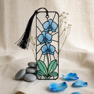 May include: A rectangular bookmark featuring a stained glass style illustration of three blue orchid flowers with green leaves. The design is outlined in black and includes a black tassel attached to the top. The bookmark has a clear, translucent finish.