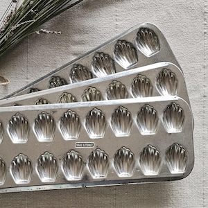 May include: Two vintage, silver-colored madeleine pans with shell-shaped molds. A bundle of dried lavender is tied with twine and rests on a neutral-colored linen surface. The pans are marked "Made in France".