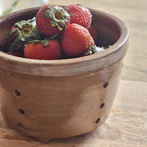 May include: A brown ceramic berry bowl with a smooth, glazed finish and small drainage holes along the sides. The bowl is filled with several fresh, bright red strawberries with green leafy stems visible at the top of the container.