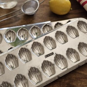 May include: A rectangular metal baking pan featuring multiple shell-shaped indentations for madeleines. The silver-colored tray has a smooth, metallic finish and includes a small rectangular label clearly marked with the text MADE IN FRANCE.