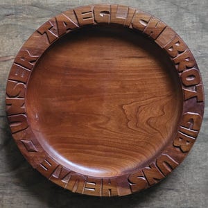 May include: A round, brown wooden plate with a smooth, polished center and a wide, raised rim. The rim features deeply carved, block-style lettering that reads "UNSER TAEGLICH BROT GIB UNS HEUTE" in a continuous circular pattern around the edge.