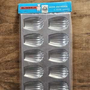 May include: A silver-colored metal madeleine baking pan with twelve shell-shaped indentations, enclosed in clear plastic packaging. The blue header card features the text BLINDALU, a chef illustration, the HOAN logo, and PETIT PATISSIER A VOTRE SERVICE.