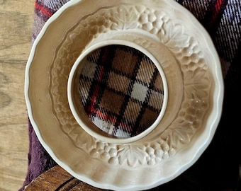 May include: A round, cream-colored ceramic saucer featuring a scalloped edge and a decorative embossed grape and leaf pattern. The center of the saucer contains a circular insert with a brown, red, and white plaid fabric pattern.