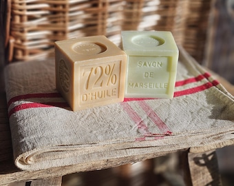 May include: Two square bars of soap rest on a linen towel with red stripes. One soap bar is beige with "72% D'HUILE" text, the other is pale green with "SAVON DE MARSEILLE" text. The background features a woven basket.
