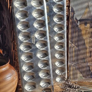 May include: Two vintage, silver-colored madeleine baking pans. Each pan has 12 shell-shaped molds. The pans are made of metal and are displayed on a wooden surface. The text "MADE IN FRANCE" is visible on one pan.