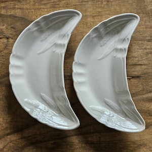 May include: Two white ceramic dishes shaped like crescent moons. Each dish features a raised, embossed wheat stalk pattern along the curved outer edge. The surface of the ceramic is smooth and glossy, with a consistent off-white color throughout.