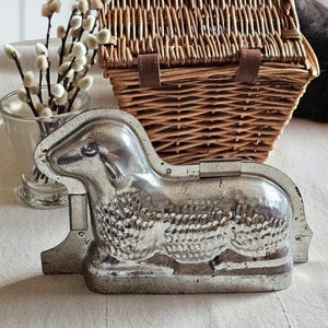 May include: A vintage, silver-colored lamb-shaped baking mold. The mold is made of metal and has detailed features. A woven wicker basket with leather straps is in the background, along with a glass vase with white pussy willow branches.