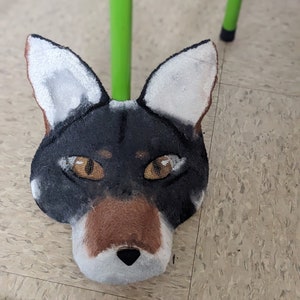 May include: A black and brown fox mask with white fur around the ears and nose. The mask has orange eyes and a black nose.