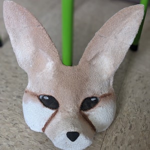 May include: A tan-colored fennec fox mask with black eyes and a black nose. The mask is made of soft, plush fabric.