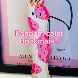 May include: A pink and white painted bookmark with a cartoon animal design. The bookmark has a white background with pink spots and a pink outline. The bookmark is titled "Random color Bookmark!!" and has the word "Picasso" written on it.