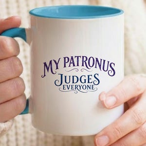 Puede incluir: Una taza de cerámica blanca con interior y asa de color azul brillante. La parte frontal de la taza presenta el texto en azul oscuro "MY PATRONUS JUDGES EVERYONE" escrito en una fuente decorativa y estilizada con elegantes adornos.