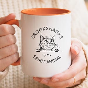 以下が含まれることがあります： オレンジ色の内側とハンドルが付いた白いセラミックマグ。マグには、猫の白黒イラストと「CROOKSHANKS IS MY SPIRIT ANIMAL」というテキストが描かれています。マグは人が持っています。