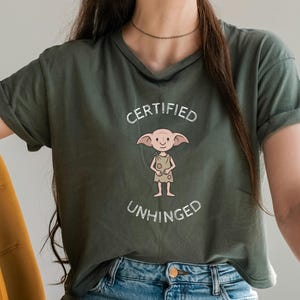 May include: Olive green t-shirt with the words "CERTIFIED UNHINGED" in white, above and below a cartoon character with large ears and a neutral expression. The shirt is made of a soft material.