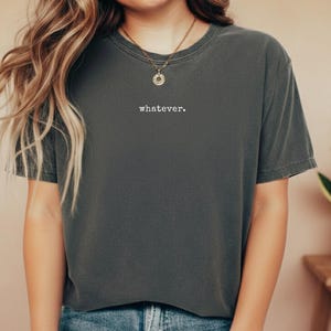 Puede incluir: Camiseta gris oscuro con la palabra "whatever." impresa en blanco. La camiseta tiene mangas cortas y cuello redondo. Se lleva un collar dorado con un colgante. La camiseta se combina con vaqueros azules.