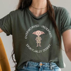 Könnte beinhalten: Olivgrünes T-Shirt mit einer Cartoonfigur und dem Text "OVERWORKED. UNDERPAID. SPIRITUALLY DOBBY." Das Design ist kreisförmig. Das Shirt hat hochgekrempelte Ärmel und einen V-Ausschnitt. Die Person trägt eine blaue Jeans.