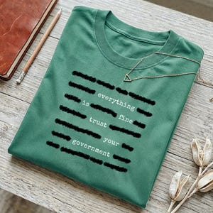 Può includere: Una maglietta verde con la scritta "everything is fine, trust your government" in bianco, parzialmente oscurata da barre nere. Una collana d'oro con un ciondolo triangolare è appoggiata sulla maglietta. Un taccuino e una matita sono sullo sfondo.