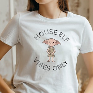 May include: White t-shirt featuring a cartoon house elf illustration with the text "HOUSE ELF VIBES ONLY" in black. The elf is depicted with large ears and a simple outfit. A casual, comfortable tee.