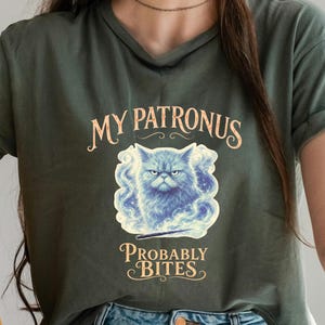 以下が含まれることがあります： 暗いオリーブグリーンのTシャツ。青い渦巻く煙と星に囲まれた不機嫌そうな猫のグラフィックが描かれています。猫の上のテキストは「MY PATRONUS」、下のテキストは「PROBABLY BITES」と書かれています。猫の下には細い黒い杖が配置されています。