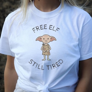 Może przedstawiać: Biała koszulka z napisem "FREE ELF STILL TIRED" w kolorze czarnym, nad i pod postacią elfa z kreskówki. Elf jest narysowany w prostym stylu, ubrany w brązowy strój. Koszulka ma klasyczny okrągły dekolt.
