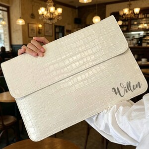 May include: Cream-colored laptop sleeve with a crocodile skin texture. The sleeve has a flap closure and the name "Willen" is printed in a cursive font on the lower right corner. The sleeve is held by a person.