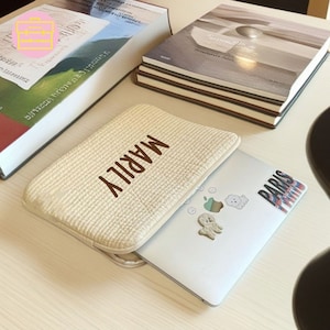 May include: A beige laptop sleeve with the name "MARILY" in brown lettering. A silver laptop with stickers is next to the sleeve. Several books are in the background.