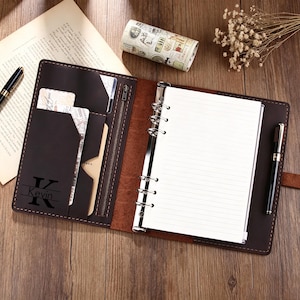 May include: Open brown leather journal with lined paper, a zippered pocket, and card slots. The journal is personalized with the name "Kevin" and the initial "K". A black pen and decorative tape roll are also visible.