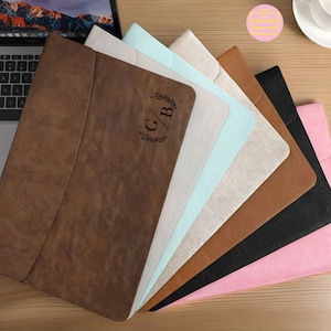 May include: A set of eight laptop sleeves in various colors, including brown, beige, light blue, brown, black, and pink. The sleeves are made of a textured material and have a flap closure. The brown sleeve has the initials "G/B" engraved on the front.