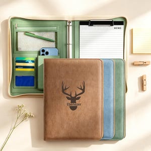 May include: A brown leather portfolio with a deer head design and the name "Ebenezer" printed on the front. The portfolio is next to blue and green portfolios. The portfolio is open, revealing a green interior with card slots, a pen, and a phone.