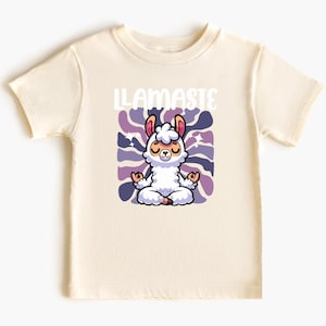 Op de afbeelding: Crème t-shirt met een cartoon lama in een yogahouding, met het woord "LLAMASTE" erboven. De lama is wit met paarse en roze accenten, tegen een paars en blauw patroon.