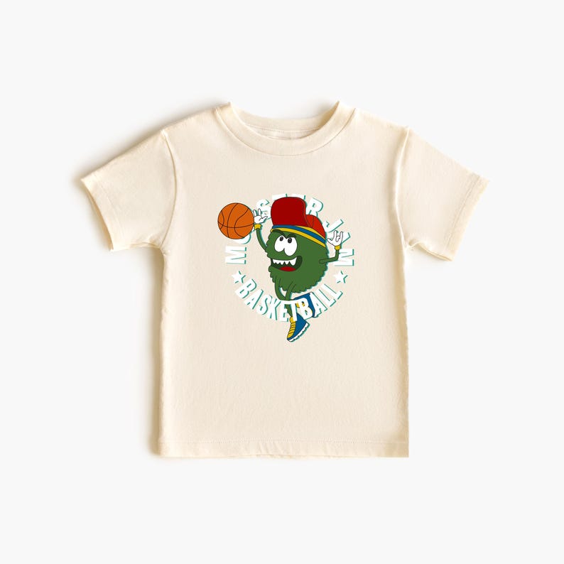 May include: A cream-coloured short-sleeved t-shirt with a cartoon character playing basketball. The character is green with a red hat and blue and yellow details. The word "BASKETBALL" is printed around the character in a circle.