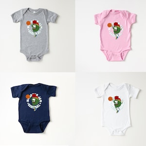 May include: Four baby onesies in grey, pink, navy blue, and white. Each onesie features a cartoon character with a basketball and the words "Monster Jam Basketball" in a circular design. The character is green with a red hat.