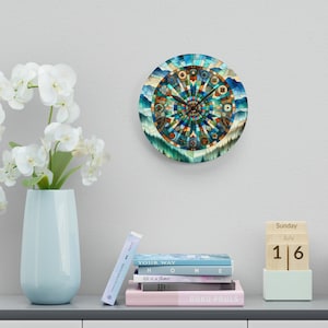 May include: A round wall clock with a blue, green, and brown mosaic design. The clock face features a circular pattern with a compass-like design. The clock hands are black and the numbers are in a circular pattern around the edge of the clock face. The clock is hanging on a white wall. The clock is surrounded by a white wall and a shelf with books and a wooden calendar.