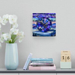 May include: A square wall clock with a stained glass design featuring a blue, purple, and white mountain landscape with a clock face in the center. The clock face is a circle with a black background and white numbers. The clock hands are black.