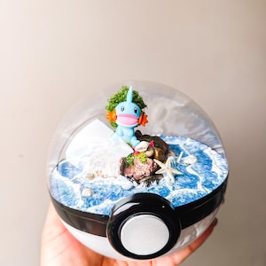 May include: A blue and orange cartoon character figurine inside a clear, spherical terrarium. The terrarium features a miniature ocean scene with a rocky island, starfish, and white waves. The base is black and white.