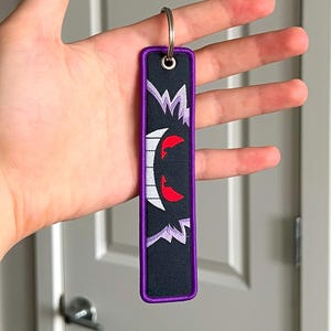Peut inclure: Porte-cl&eacute;s rectangulaire noir avec une bordure violette et un anneau argent&eacute;. Le porte-cl&eacute;s pr&eacute;sente un motif de visage de fant&ocirc;me de dessin anim&eacute; blanc et rouge. Le motif est brod&eacute; sur le tissu noir.
