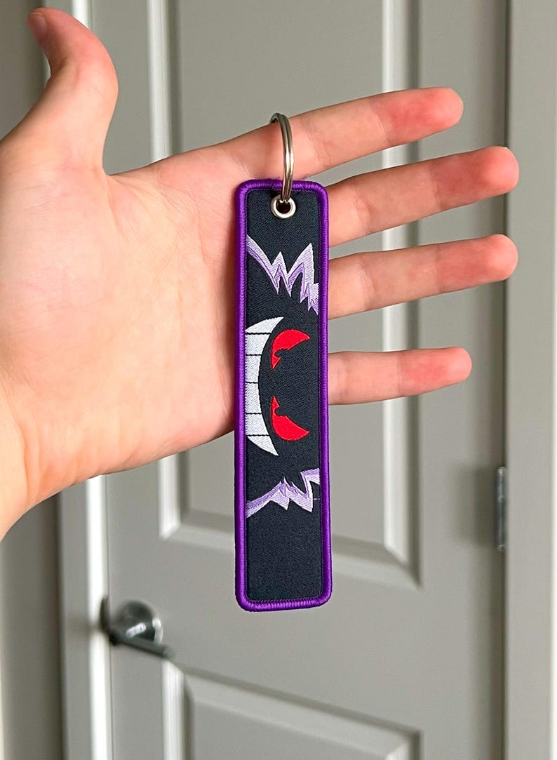 Peut inclure: Porte-cl&eacute;s rectangulaire noir avec une bordure violette et un anneau argent&eacute;. Le porte-cl&eacute;s pr&eacute;sente un motif de visage de fant&ocirc;me de dessin anim&eacute; blanc et rouge. Le motif est brod&eacute; sur le tissu noir.