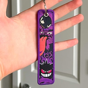 Peut inclure: Un porte-cl&eacute;s brod&eacute; violet et noir repr&eacute;sentant trois personnages de fant&ocirc;mes de dessins anim&eacute;s. Le porte-cl&eacute;s a un anneau m&eacute;tallique en haut pour attacher les cl&eacute;s. Le design comprend un fant&ocirc;me noir avec une bombe, un fant&ocirc;me violet avec une langue rouge et un fant&ocirc;me noir avec des yeux rouges.