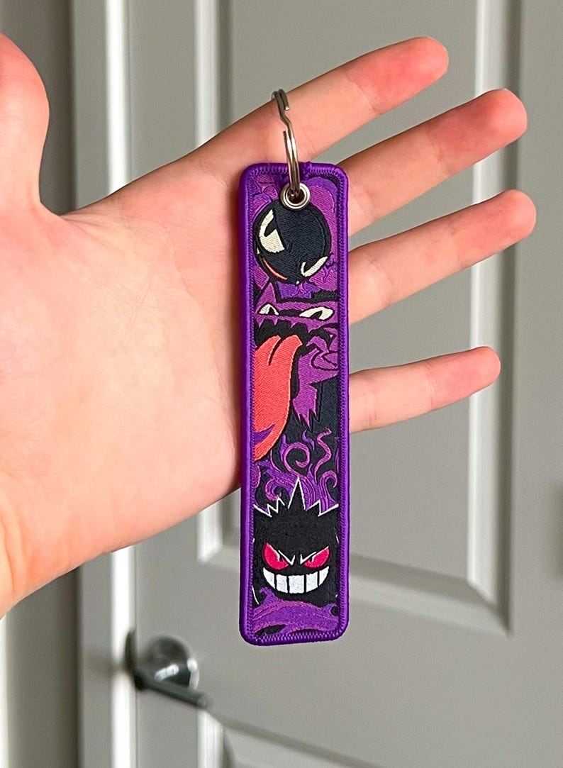 Peut inclure: Un porte-cl&eacute;s brod&eacute; violet et noir repr&eacute;sentant trois personnages de fant&ocirc;mes de dessins anim&eacute;s. Le porte-cl&eacute;s a un anneau m&eacute;tallique en haut pour attacher les cl&eacute;s. Le design comprend un fant&ocirc;me noir avec une bombe, un fant&ocirc;me violet avec une langue rouge et un fant&ocirc;me noir avec des yeux rouges.