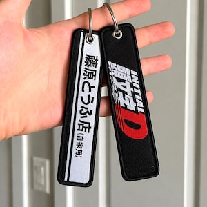 May include: Two rectangular fabric keychains with metal rings. One is white with black text reading 藤原とうふ店(自家用) and a black border. The other is black with white text reading INITIAL D and a large red letter D. Both feature embroidered edges.